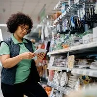 A woman in a green shirt and vest uses a tablet while standing in a store aisle filled with various products.